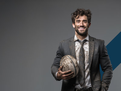 A photo of a muddy businessman holding a rugby ball and smiling